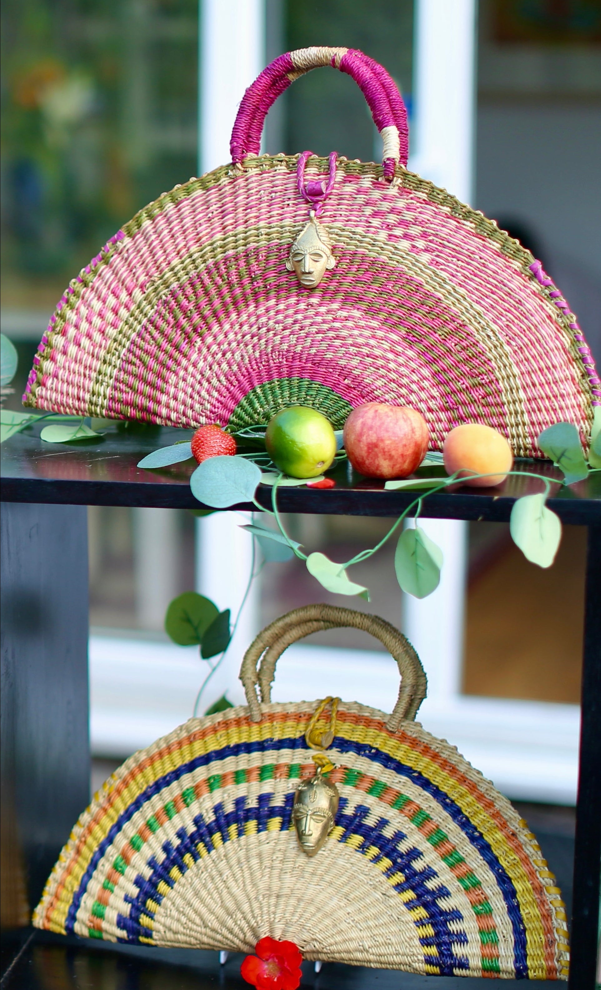 Two colorful woven handbags on a glass table with fruits and leaves.