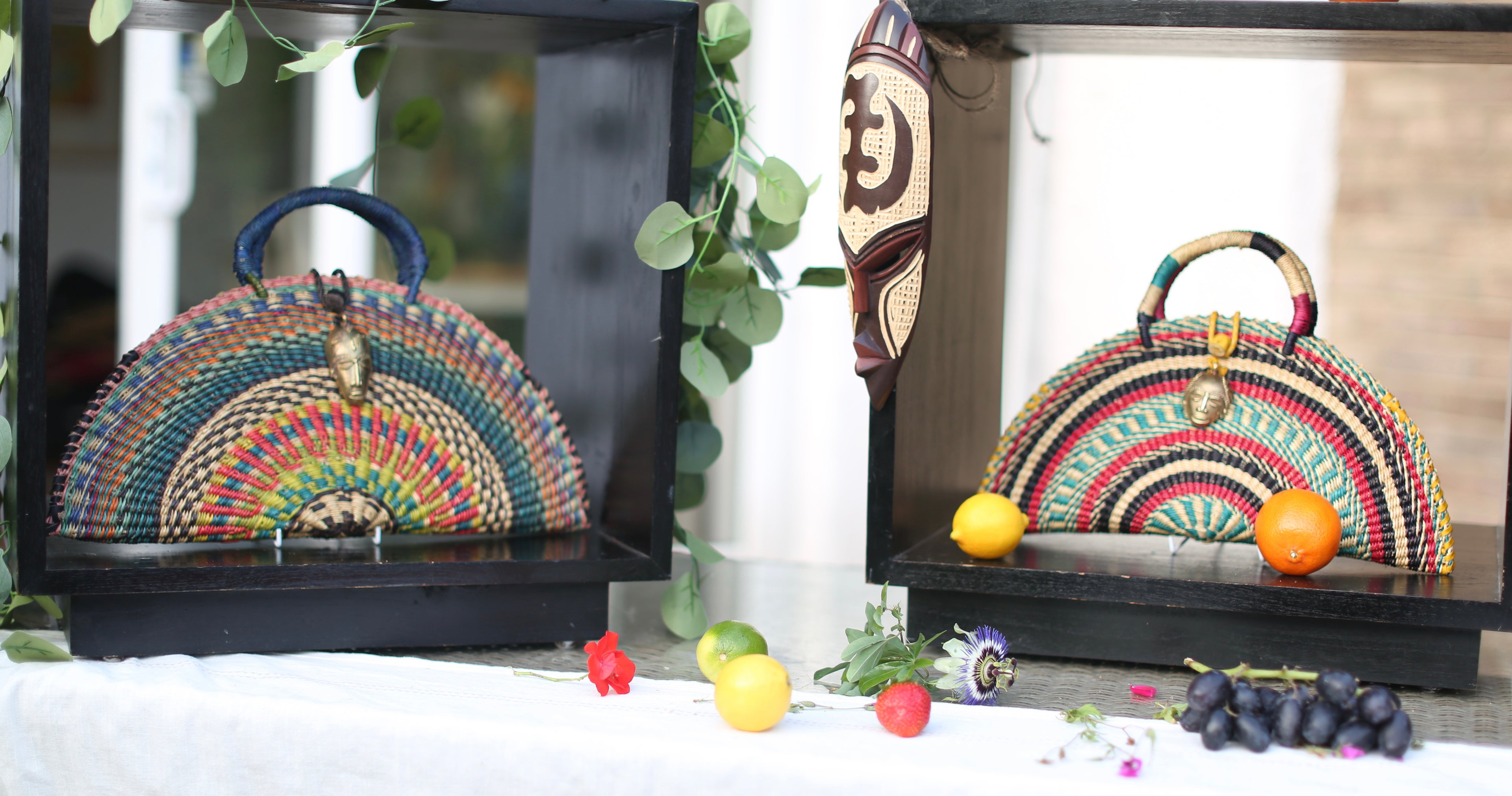 Two colorful handbags on display with fruits and leaves in the foreground.